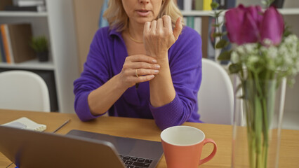 A mature caucasian woman in a purple sweater checks her wrist at home, suggesting joint pain or time.