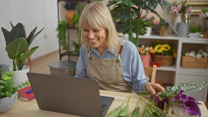 Smiling blonde woman in apron using laptop among vibrant flowers inside a florist shop.