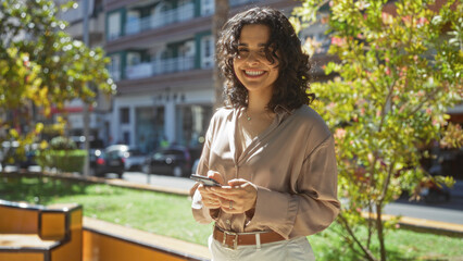 A beautiful brunette hispanic woman smiling outdoors in an urban park setting, holding a smartphone in a city environment on a sunny day.