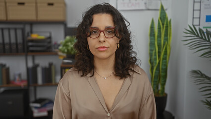 Hispanic woman with brunette hair and glasses in an office room, featuring a workplace setting with bookshelves and plants indoors, looking confidently at the camera.