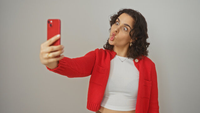 A young, hispanic, attractive woman with brunette hair taking a playful selfie against a white background, wearing a red sweater and a white crop top, making a funny face.