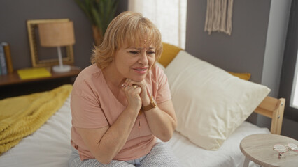 A middle-aged caucasian woman with blonde hair sits on a bed in her bedroom, appearing thoughtful or concerned, with indoor home decor visible in the background.