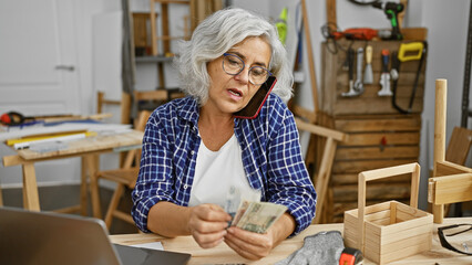 A mature woman counts polish zloty bills in a carpentry workshop while talking on a smartphone.