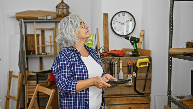 Mature woman in workshop inspecting work with tablet amidst carpentry tools