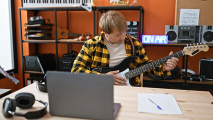 A young caucasian man plays an electric guitar in a music studio with recording equipment and 'on...