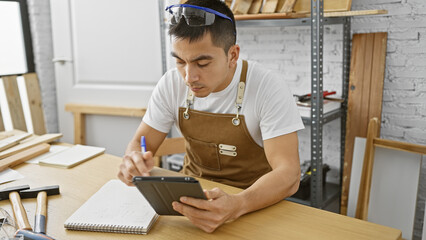 A handsome young hispanic man uses a tablet for planning in a well-equipped carpentry workshop indoors.