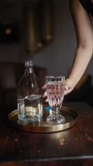 A woman serves water from a clear bottle into a glass on a brass tray, illustrating hospitality or service.