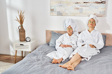 Obraz premium A brunette mother and daughter in white bath robes sitting on a bed, with towels wrapped around their heads.