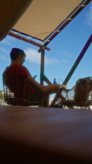 Man relaxing under a tent in african nature with a phone in hand, surrounded by adventure and camping gear.