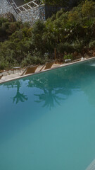 A serene outdoor pool bordered by lush greenery and stone wall reflects palm trees under a bright sky, with empty lounge chairs beside for relaxation.