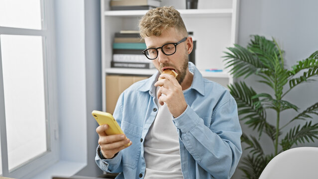Bearded man in glasses eats a sandwich while checking his smartphone in a modern office. - Powered by Adobe