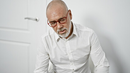 Obraz premium Pensive senior man with glasses and beard in white shirt indoors