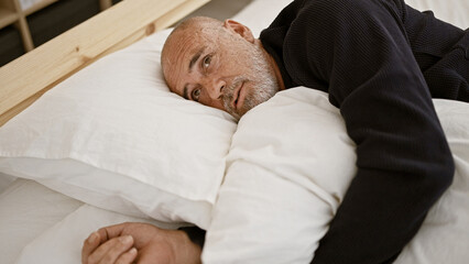 Mature bald man with beard lying in bed looking thoughtful in a bedroom setting, portraying a sense...