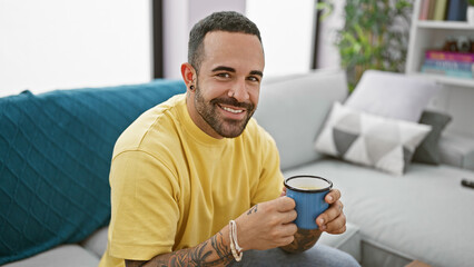 Smiling young hispanic man with a beard holding a coffee cup in a cozy living room.