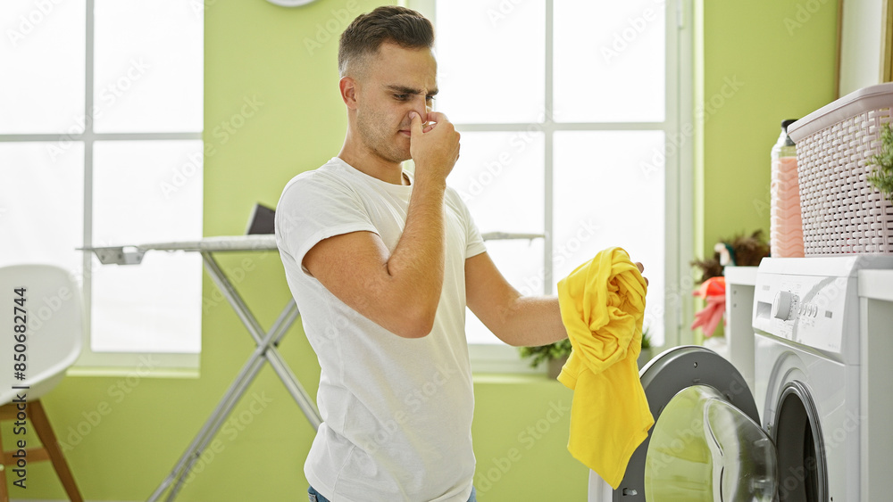 Wall mural a young man grimaces at a foul-smelling shirt in a bright laundry room.