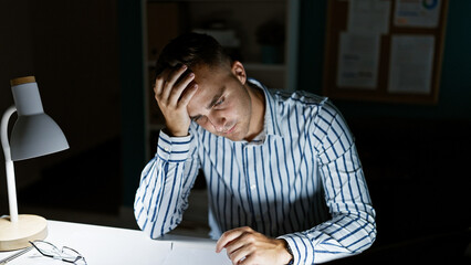 A stressed young man sits indoors at a dimly lit office desk, showing frustration as he reads...