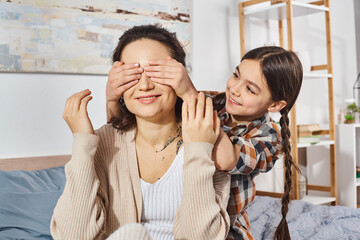A girl covering eyes of her mother, sharing a playful moment together at home.