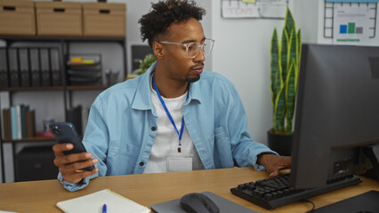 Handsome young african american man with a beard working in an office environment, using a computer and holding a mobile phone, with shelves and plants in the background.