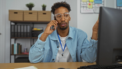 Handsome young african american man with beard in an office room wearing eyeglasses and a blue shirt, speaking on a phone call while sitting at a desk with computer screen in front.