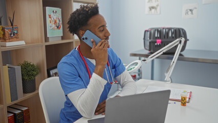 A handsome young african american male vet, wearing blue scrubs and a stethoscope, talks on the phone in a well-organized vet clinic office with books, plants, and a laptop on the desk.