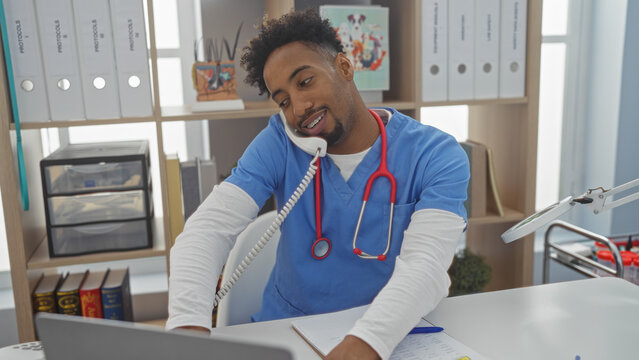 Handsome african american man with a beard working in a veterinary clinic, wearing scrubs and a stethoscope, multitasking with a phone and computer in a bright, organized interior.
