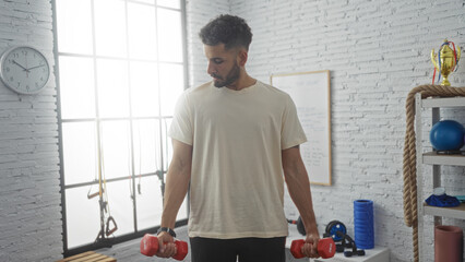 A young, hispanic man lifting dumbbells in a brightly lit gymnasium with fitness equipment and trophies in the background.