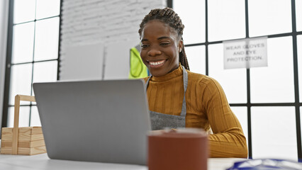 A smiling african american woman with braids wearing an apron uses a laptop in a bright carpentry...