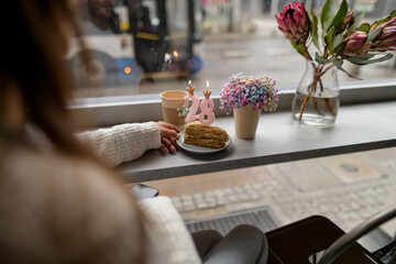 Woman in front of the window with a glass of coffee, birthday cake and flowers