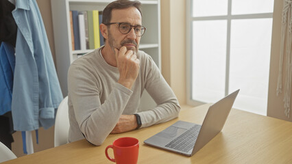 A thoughtful senior man with glasses using a laptop at a home office, a red mug on the table and daylight illuminating the room.