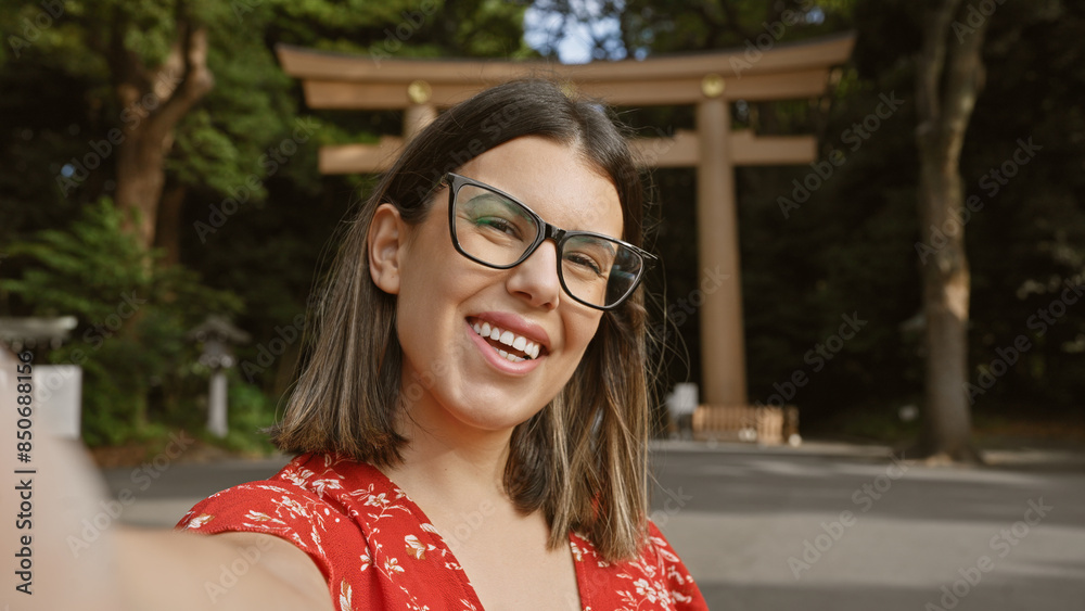 Poster Cheerful, beautiful hispanic woman wearing glasses having fun making a confident selfie picture, smiling expression at meiji temple, tokyo's cultural shrine, capturing her happy moment.