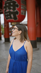 Cheerful, beautiful hispanic woman with glasses joyfully exploring and admiring the majestic senso-ji temple as she casually strolls, looking around with awe and a confident smile.