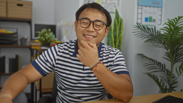 Handsome young chinese man with glasses smiling confidently in a modern office room with indoor plants and bookshelf.
