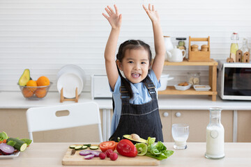 cute asian little child celebrating her success with raised arms pose after drinking milk and eating vegetables in the kitchen