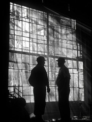 Silhouettes of two men standing in front of large industrial windows, with light streaming through in a dramatic effect.