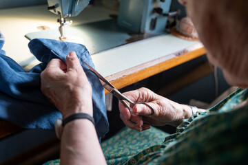 chubby eighty-year-old woman in green summer dressing gown mending and sewing the bottom of a pair of jeans on the sewing machine.