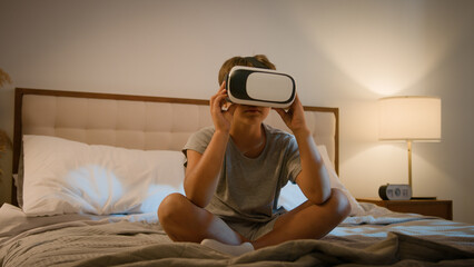 Little modern Caucasian child wearing vr glasses virtual reality headset boy son bed playing online 3d virtual game kid exploring digital universe world game alone evening illuminated bedroom indoors