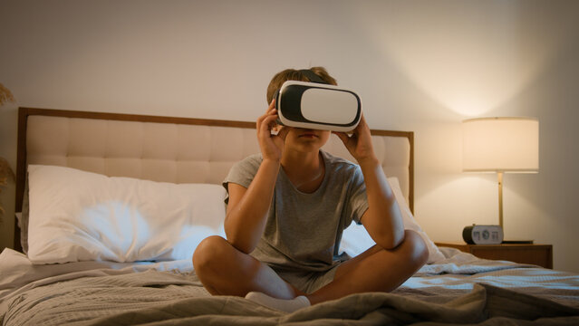 Little modern Caucasian child wearing vr glasses virtual reality headset boy son bed playing online 3d virtual game kid exploring digital universe world game alone evening illuminated bedroom indoors