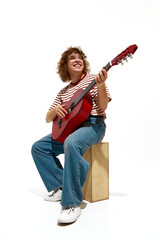 Fototapeta premium Portrait of happy young man, musician wearing casual clothes, sitting on wooden box and playing guitar isolated on white background. Concept of music, festival, concert, hobby, talent, performance