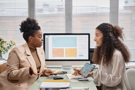 Side view of two businesswomen discussing new mobile app interface design while holding color swatches. Diverse team talk shop against window in office, copy space