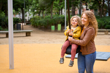 Mother and daughter are at a park, with the woman holding the child on a swing