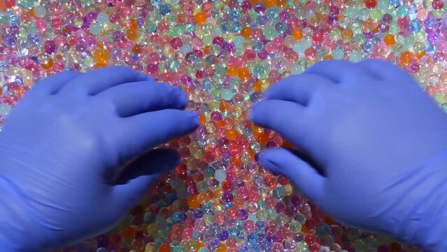 Hands in blue gloves smoothly sink into a multitude of multicolored orbis. The hands are moving in a lot of colored, shiny orbis. View from above. ASMR Part13.14