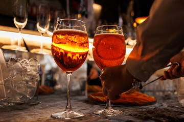 Bartender makes two glasses of cocktail Aperol spritz on bar counter, adds fresh orange slices. Typical alcoholic Italian beverage, aperitif made with Prosecco sparkling white wine and ice cubes.