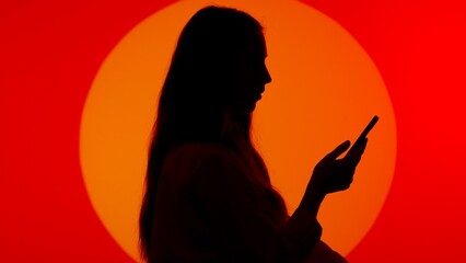 Stylish young woman black silhouette in casual using smartphone, scrolling social media online. Isolated on red studio background spotlight.