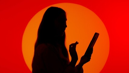 Stylish young woman black silhouette in casual using tablet, scrolling social media online. Isolated on red studio background spotlight.
