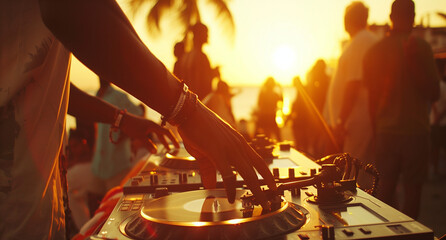 A close-up shot of hands on turntables, with people dancing in soft focus behind at sunset during an outdoor party. A photo of DJ equipment on the beach with a sunset and crowd in the background. 