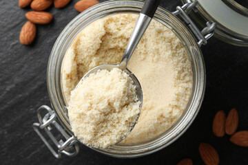 Spoon with fresh almond flour above jar and nuts on dark textured table, top view