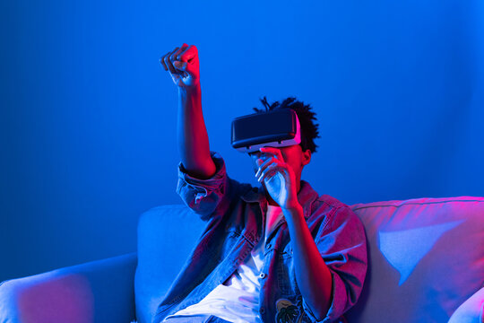 Young African American looking VR enjoy watching fantastic 3D pointing interesting object technology blue neon light virtual reality metaverse new world at living room sitting on sofa. Contrivance.