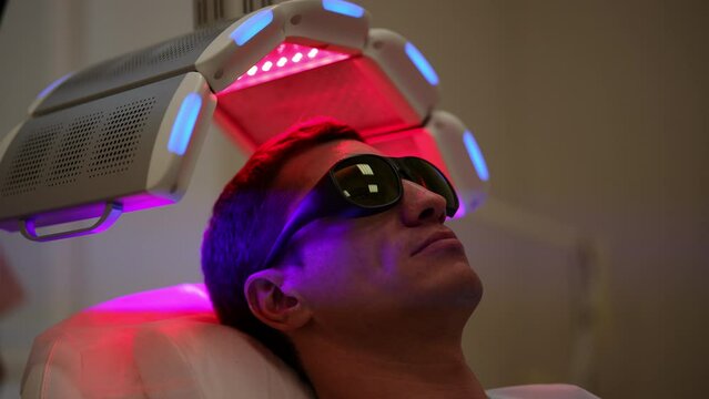 Closeup face of handsome young man having LED light hair photodynamic therapy treatment after hair transplant at clinic. Male client having non-invasive type of phototherapy. Shooting in slow motion.