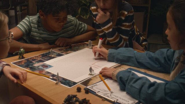 Side shot of girl drawing location map with red marker pen while playing table game with diverse friends at home in evening