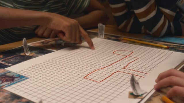 Closeup of hands of unrecognizable children explaining how to draw location map to friend who using red marker pen while playing board game together indoors